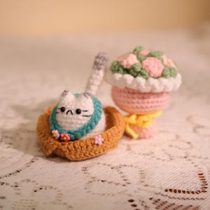 May include: Crocheted cat figurine in a brown basket, wearing a teal scarf and adorned with pink flowers. A crocheted pink flower pot with a white rim and a bouquet of pink and green flowers is also present. The items are handmade.