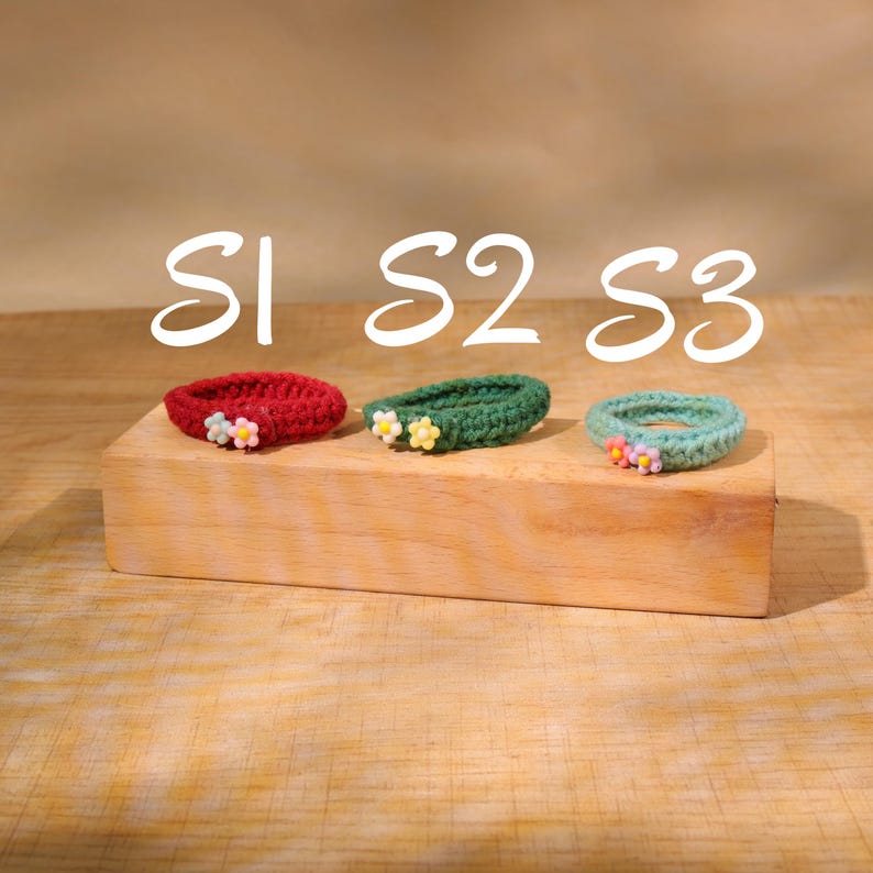 May include: Three crocheted hair ties in red, green, and teal, each adorned with small, colorful flower accents. The hair ties are displayed on a wooden block, with the text "S1 S2 S3" above them.