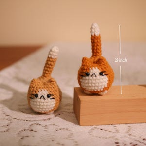 May include: Two handmade crochet cat figurines. The cats are orange and white, with black facial details and a tail that is 3 inches long. The figurines are sitting on a wooden block.