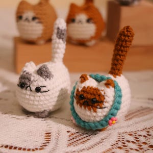 May include: Crocheted cat figurines in white, gray, and brown. One cat has a gray and white pattern, while another has a brown and white face with a teal collar and a small pink flower. Other cat figurines are in the background.