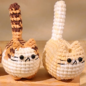 May include: Two handmade crochet cat figurines. One cat is cream and orange with a striped tail, the other is cream and yellow with a white tail. Both have black bead eyes and are sitting upright.