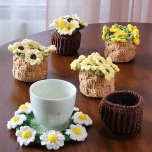 Può includere: Una collezione di articoli all'uncinetto fatti a mano, tra cui piccoli coprivasi con fiori gialli e bianchi, un sottobicchiere a margherita e una tazza da tè. L'uncinetto presenta un motivo a trama di cestino.