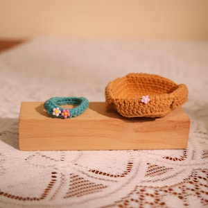 May include: A teal crocheted bracelet with pink and yellow flower accents sits next to a small, tan crocheted basket. Both items are displayed on a wooden block, set against a soft, neutral background.