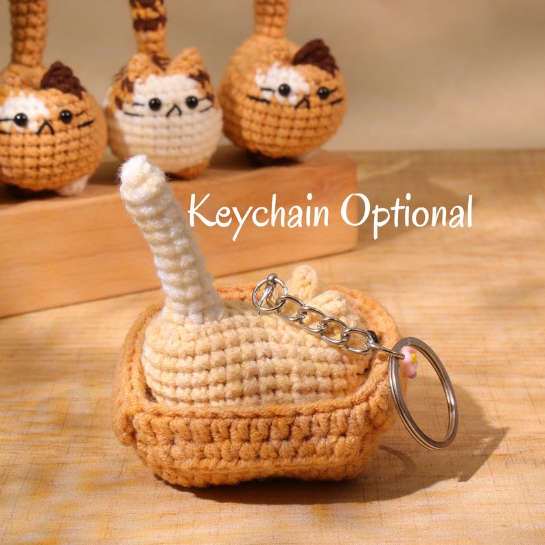May include: Crocheted cat keychain in shades of tan, cream, and brown. The keychain features a small, round cat with a tail and a metal keyring. The text "Keychain Optional" is visible in the image. Other similar cat keychains are in the background.