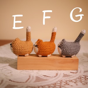 May include: Three crocheted cat figurines in shades of orange, tan, and gray, each with a white belly and tail tip. The cats are positioned on a wooden block, with the letters "E", "F", and "G" above them.