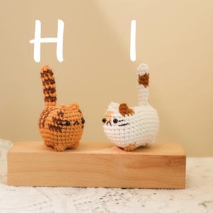 May include: Two crocheted cat figurines, one orange tabby and one white with brown patches, stand on a wooden block. The word "HI" is written above them in white. The cats have black eyes and upright tails.