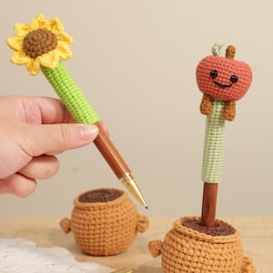 May include: Crocheted pens with whimsical designs. One pen features a sunflower with yellow petals and a brown center, while the other has a smiling red apple. Both pens have green stems and are set in small, brown crocheted pots.