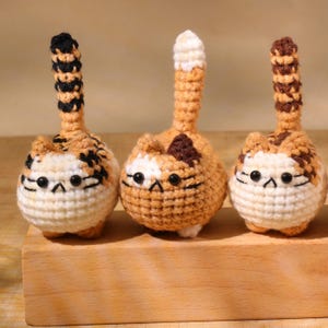 May include: Three handmade crochet cat figurines. Each cat has a round body, a long tail, and small black eyes. The cats are primarily orange, white, and brown, with varying patterns on their bodies and tails. They are displayed on a wooden block.