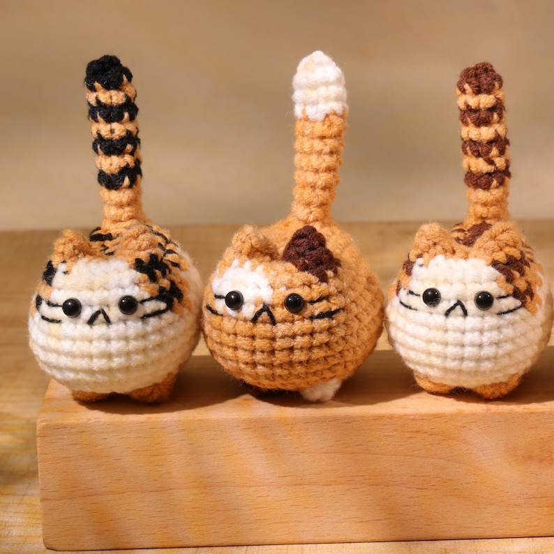 May include: Three handmade crochet cat figurines. Each cat has a round body, a long tail, and small black eyes. The cats are primarily orange, white, and brown, with varying patterns on their bodies and tails. They are displayed on a wooden block.
