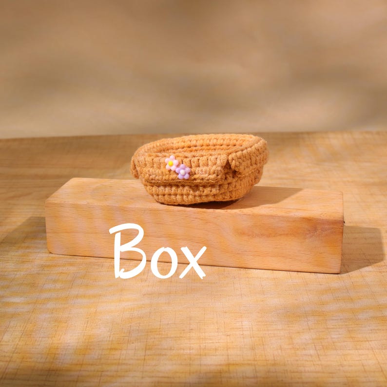 May include: A small, crocheted, tan-colored basket with a decorative flower. The basket is sitting on a wooden block with the word "Box" written on it. The background is a light brown color.