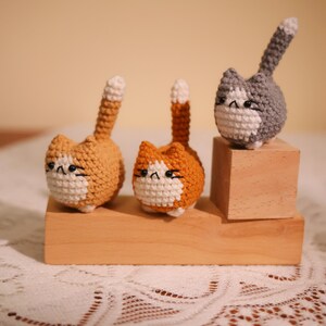 May include: Three crocheted cat figurines in shades of orange, gray, and white. Each cat has a distinct expression and a raised tail. They are displayed on a wooden block, showcasing the handmade craftsmanship. The cats are a charming addition to any cat lover's collection.