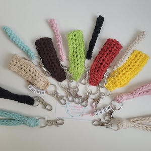 May include: A collection of colorful crocheted keychains with different patterns and designs. Each keychain has a metal ring and a small charm with the word "ROSA" or "MAIA" on it.