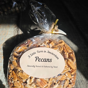 Könnte beinhalten: Ein klarer Plastikbeutel gefüllt mit Pekannusshälften, gebunden mit Bindfaden und einem goldenen Band. Das Etikett lautet "A Little Farm in Amsterdam Pecans Naturally Raised & Gathered by Hand."
