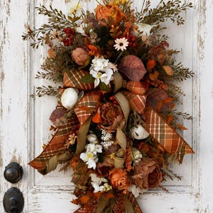 May include: A teardrop-shaped floral swag featuring orange, rust, and white artificial flowers, green foliage, and small red berries. The arrangement is accented with a wide plaid ribbon in shades of orange, brown, and green, tied in a bow.