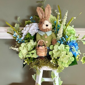 May include: An Easter-themed decorative arrangement featuring a woven bunny wearing a green sweater and holding a basket of eggs. The bunny is surrounded by green, blue, and white faux flowers and foliage, displayed on a white, three-tiered stand.