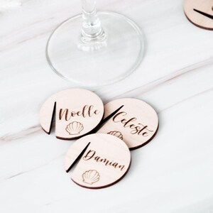 May include: Wooden wine glass charms with names "Noelle", "Celeste", and "Damian" engraved in a script font. Each charm has a seashell design. The charms are light brown and are placed on a white marble surface.
