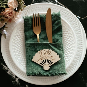 May include: A white plate with a green napkin and gold silverware. The napkin is folded and has a wooden fan-shaped place card with the name "Katie" written on it.