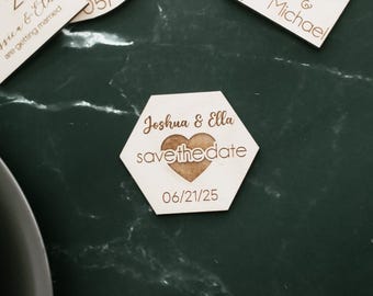 May include: Wooden hexagon-shaped save-the-date magnet with the names "Joshua & Ella" and the date "06/21/25" engraved. A heart shape is cut out in the center. Other wooden magnets are visible in the background.