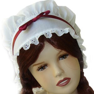 May include: White lace bonnet with a burgundy ribbon and bow. The bonnet has a ruffled edge and is worn on a mannequin with long, dark brown hair.