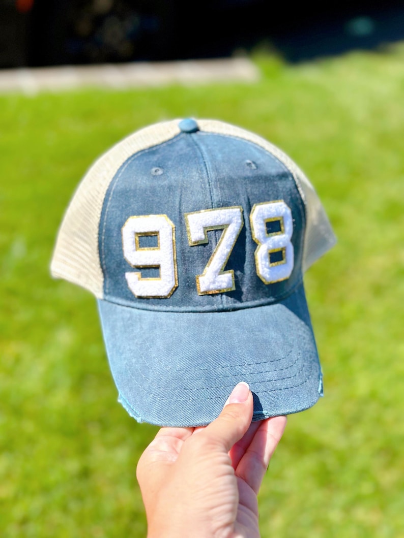 K&ouml;nnte beinhalten: Eine blaue und wei&szlig;e Trucker-Kappe mit der Nummer "978" in Wei&szlig; mit goldener Umrandung auf der Vorderseite.