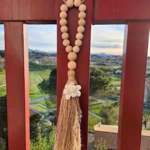 Può includere: Una ghirlanda di perline di legno con un fiocco bianco e un ciondolo a forma di fiore bianco. La ghirlanda è appesa a una recinzione di legno rosso.