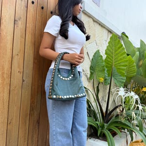 May include: A dark green hobo-style handbag featuring a curved top handle and a structured base. The bag is adorned with a series of metallic, circular studs lining the edges of the handle and the bottom perimeter of the bag's main body.