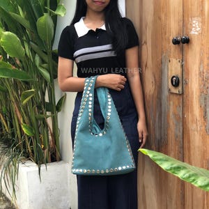 May include: Teal suede tote bag with silver-colored stud accents. The bag has a unique triangular shape and a single shoulder strap. The text "WAHYU LEATHER" is visible on the image.