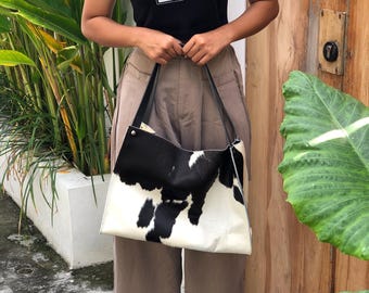 May include: A large tote bag with a cowhide pattern in black and white is held. The bag has black straps. The person is wearing a black t-shirt with the number 28 in white and tan trousers.