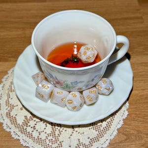 May include: A white teacup and saucer set with floral patterns, containing a liquid and a 20-sided die. Additional dice are arranged on the saucer. The teacup rests on a lace doily on a wooden surface. Ideal for tabletop gamers.