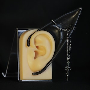 Puede incluir: Un pendiente de oreja plateado con un dije de espada colgante se muestra en una forma negra con forma de oreja. La forma de la oreja está montada en un soporte de exhibición transparente, sobre un fondo negro. La oreja es de color beige claro.