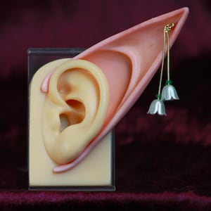 May include: A pair of elf ears with a delicate flower earring. The earring features a white flower with a green stem and a silver bead.