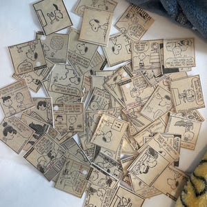 May include: A collection of vintage Peanuts comic strips featuring Snoopy and Charlie Brown. The strips are printed on small, rectangular pieces of paper with a tan background. The strips are scattered on a white surface.