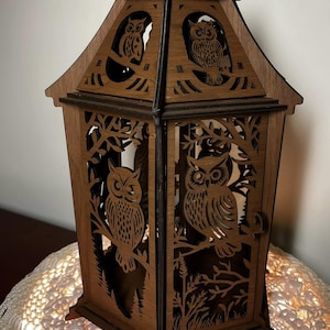 May include: A wooden lantern with a hexagonal base and a handle on top. The lantern is decorated with intricate cutouts of owls and branches. The lantern is lit from within, casting a warm glow.