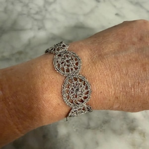 May include: A metallic silver bracelet featuring a series of interconnected circular crochet motifs. The design consists of three distinct, round, lace-like patterns linked together to form a continuous band that wraps around the wrist.