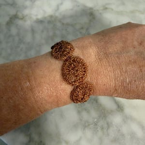 May include: A copper-coloured bracelet featuring three circular, interconnected wire-crocheted medallions. The metallic wire is woven into a delicate, open-work pattern, creating a series of linked, round shapes that form the band of the bracelet.