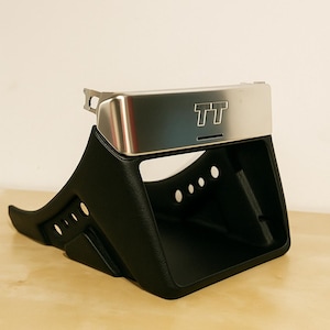Può includere: Un componente automobilistico nero con un accento argentato con le lettere "TT". Il componente ha un design curvo con diverse aperture circolari ed è posto su una superficie di legno di colore chiaro.