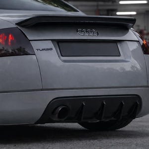 Può includere: Vista posteriore di un'auto sportiva Audi TT argentata. Le caratteristiche includono uno spoiler nero, un diffusore posteriore nero e un singolo tubo di scarico. La parola "TURBO" è visibile. Fanali posteriori rossi illuminati.