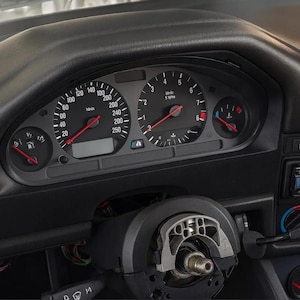 Könnte beinhalten: Nahaufnahme eines schwarzen Armaturenbretts mit Tachometer und Drehzahlmesser. Der Tacho zeigt bis zu 250 mph an, der Drehzahlmesser bis zu 8 U/min. Das Lenkrad ist teilweise sichtbar. Das Armaturenbrett ist schwarz.
