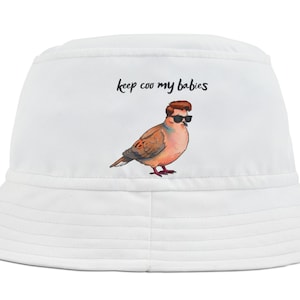 May include: White bucket hat with a cartoon bird wearing sunglasses and a brown toupee. The text "keep coo my babies" is above the bird. The hat has a wide brim and is made of a soft material.