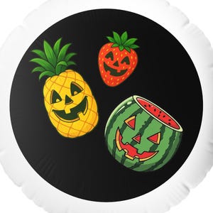 Op de afbeelding: Een ronde witte ballon met een zwart middenstuk met cartoon pompoengezichten op een ananas, een aardbei en een watermeloen. De ananas is geel, de aardbei is rood en de watermeloen is groen.