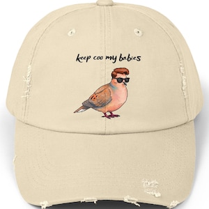 May include: A distressed beige baseball cap featuring a cartoon dove wearing sunglasses. The text "keep coo my babies" is above the bird. The cap has a curved brim and a distressed look.
