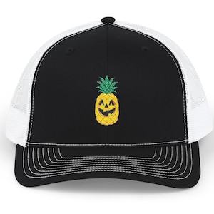 May include: Black and white trucker hat with a smiling jack-o'-lantern pineapple design. The hat features a black front panel with the embroidered pineapple, a white mesh back, and a curved brim with white stitching. A fun accessory for Halloween.