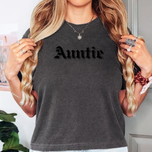 Pode incluir: Uma camiseta cinza escura cortada com a palavra "Auntie" impressa em letras góticas pretas.