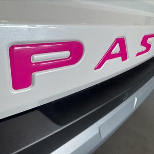 May include: Close-up of a car's rear bumper with the word "PASS" in bright pink, glossy, raised lettering. The bumper is white with a black and silver lower section. The letters are bold and stand out.
