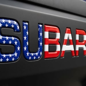 Pode incluir: A palavra SUBARU em letras tridimensionais em relevo sobre uma superfície preta fosca. As letras S, U e B apresentam um fundo azul com estrelas brancas, enquanto as letras A, R e U exibem listras horizontais vermelhas e brancas.