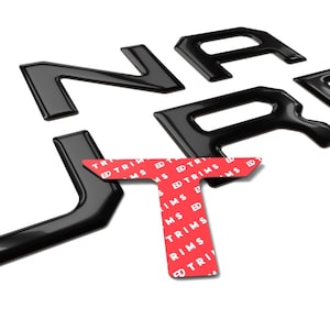 May include: Black automotive lettering spelling out "UNARO", with a red and white sticker that says "TRIMS". The letters are glossy and appear to be made of plastic or metal. The letters are arranged on a white background.