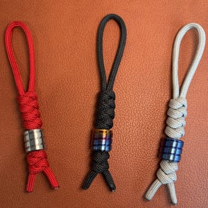 May include: Three keychains with braided cord loops. One is red with a silver bead, another is black with a gold and blue bead, and the third is gray with a blue bead. The keychains are displayed on a brown surface.