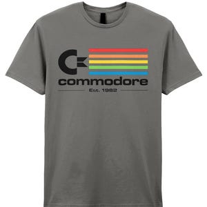 Könnte beinhalten: Ein graues T-Shirt mit dem Commodore-Logo in Schwarz und einem Regenbogenstreifen-Design. Das Wort "Commodore" ist unter dem Logo aufgedruckt, darunter "Est. 1982". Das T-Shirt hat einen Rundhalsausschnitt und kurze Ärmel.