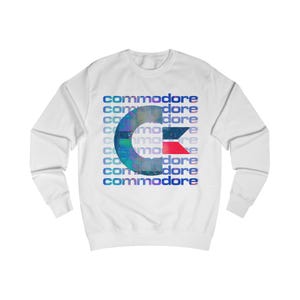 May include: White sweatshirt featuring the word "commodore" in a repeating pattern. A large, stylized "C" in shades of blue, green, and red is centered on the front. The sweatshirt has long sleeves and a crew neck.
