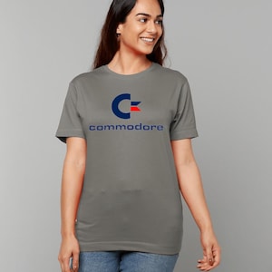 Könnte beinhalten: Ein graues T-Shirt mit einem blauen und roten Commodore-Logo und dem Wort "commodore" in blau unter dem Logo.